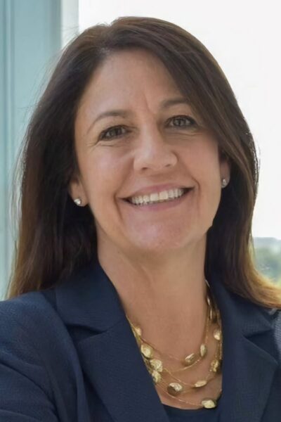 A smiling woman with shoulder-length dark hair wearing a dark blazer and a gold necklace, standing indoors near a large window with a view of greenery outside.